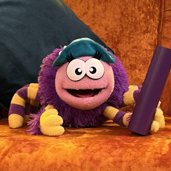 Tinsy Spider Playtime Towers. A Purple Spider Puppet with a blue turquoise hat holding a cardboard tube. She has yellow and purple striped legs. Puppet Design Puppet Manufacture by Talk to the Hand Puppets